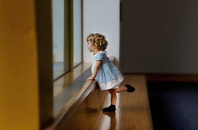 Dievčatko v modrých šatách stojí pri veľkom okne